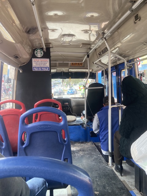 Das Bild zeigt das innere eines Busses in Cusco. Zu sehen sind rote und blaue Sitze, drei Personen und die Windschutzscheibe.