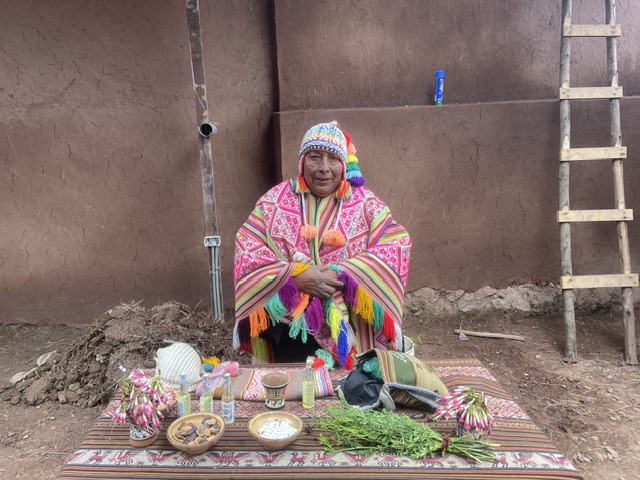 Das Bild zeigt einen Schamanen der Quechua in seiner Tracht mit Poncho und Andenmütze. Seine Kleidung ist rot-weiß mit bunten Ornamenten. Vor ihm befindet sich ein Tisch mit verschiedenen Utensilien für ein Pachamama-Ritual, einem Ritual für die Mutter Erde. Dazu gehören Kokablätter, Duftwässer und Kräuter.