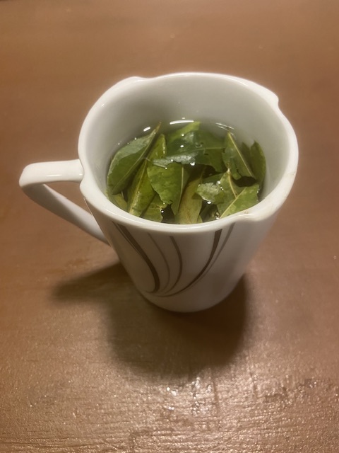 Das Bild zeigt eine Tasse Mate de Coca - Kokatee. Zu sehen sind die grünen Blätter in heißem Wasser in einer weißen Tasse mit schwarzen Linien.