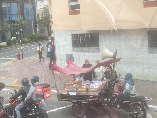 Das Bild zeigt einen Orangenverkäufer auf seinem Verkaufswagen in einer Straße von Cusco. Es hat vorne zwei Räder, worüber sich seine Ware befindet. Darüber wurde ein rotes Tuch als Sonnenschutz befestigt.