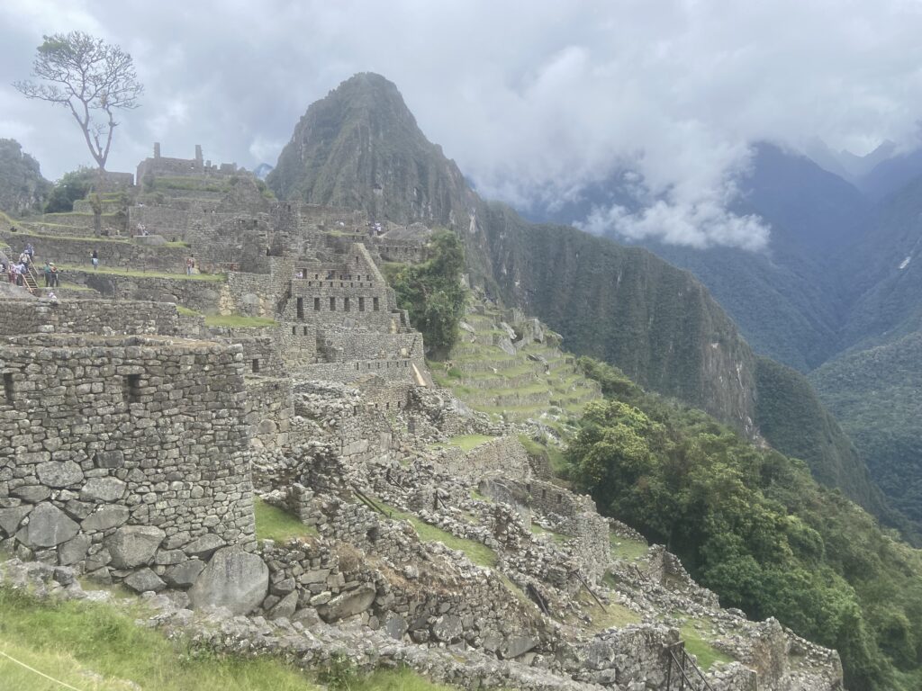 Das Bild zeigt einen Teil der archäologischen Stätte Machu Picchu. Darauf sind einige Ruinen und im Hintergrund der Huayna Picchu zu sehen.