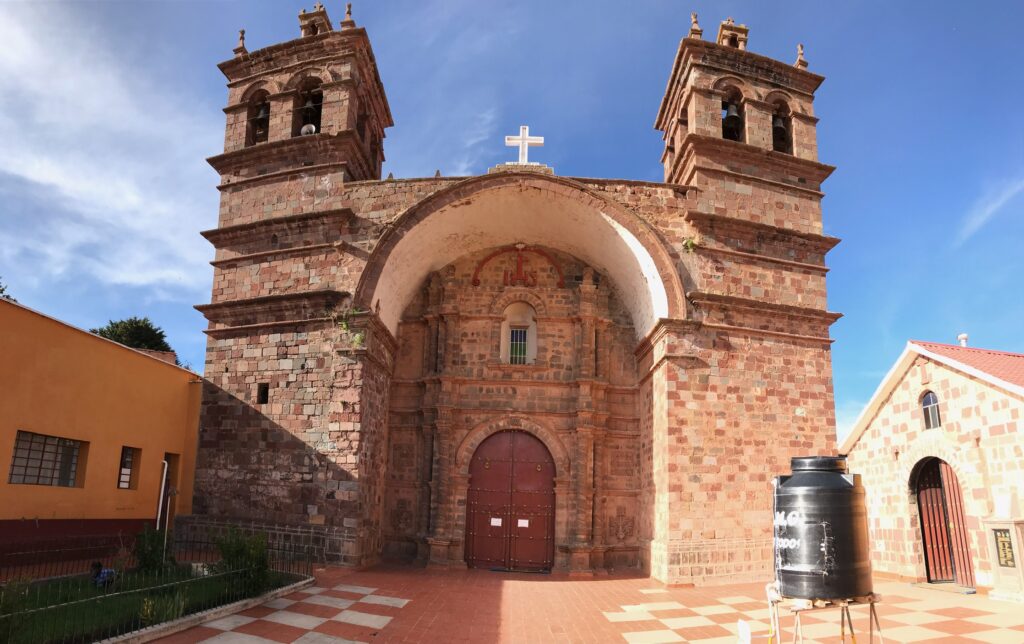 Das Bild zeigt die Kirche von Guaqui auf dem Altiplano in der Nähe des Titicacasees. Das Gebäude besteht aus hellen beigen und braunen Steinen und hat auf der Frontseite zwei Glockentürme - in der Mitte ist ein kleines weißes Kreuz zu sehen. Vor der Kirche steht ein schwarzer Wassertank.