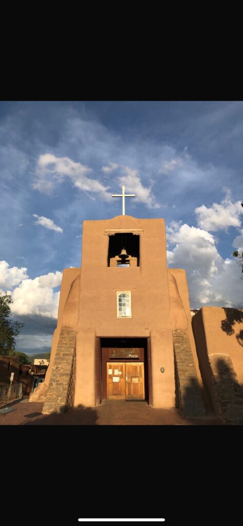 Das Bild zeigt die Missionskirche San Miguel in Santa Fe (New Mexico, USA) und gilt als älteste erhaltene Kirche nördlich von Mexiko. Sie ist beigefarben, hat einen kleinen Glockenturm und eine hölzerne Tür. Auf dem Glockenturm befindet sich ein weißes Kreuz. Im 17. Jahrhundert erbaut auf Überresten von vorspanischen Gebäudestrukturen steht sie südlich des von den Spaniern erbauten Zentrums in einem Viertel, das ursprünglich von Tlaxkalteken, indigenen Hilfstruppen der Spanier erbaut wurde.