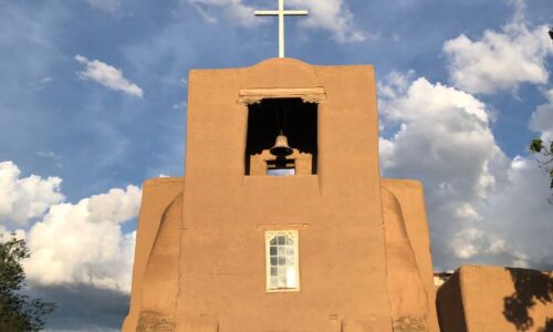 Das Bild zeigt die Missionskirche San Miguel in Santa Fe (New Mexico, USA) und gilt als älteste erhaltene Kirche nördlich von Mexiko. Sie ist beigefarben, hat einen kleinen Glockenturm und eine hölzerne Tür. Auf dem Glockenturm befindet sich ein weißes Kreuz. Im 17. Jahrhundert erbaut auf Überresten von vorspanischen Gebäudestrukturen steht sie südlich des von den Spaniern erbauten Zentrums in einem Viertel, das ursprünglich von Tlaxkalteken, indigenen Hilfstruppen der Spanier erbaut wurde.